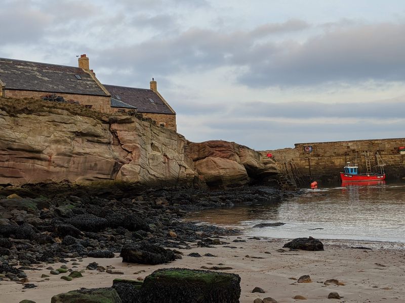 Stone cottages built against layered red cliffs beside the rocky shore at Cove Harbour, with a small red boat in the water.
