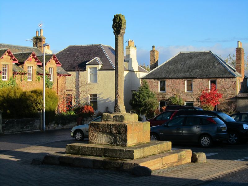 The tall stone mercat cross on its stepped base in Cockburnspath square, surrounded by houses and autumn trees.